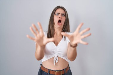 Gündelik beyaz tişört giyen genç ve güzel bir kadın korku ifadesi yüzünden korkmuş ve dehşete düşmüş. El hareketlerini bırak, şok içinde bağır. panik konsepti. 