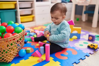 Adorable hispanic baby playing with construction blocks sitting on floor at kindergarten