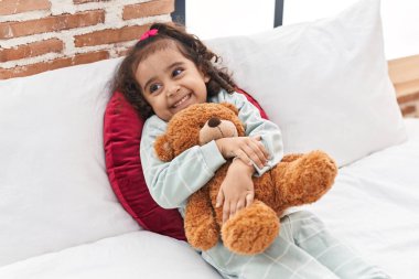 Adorable hispanic girl hugging teddy bear lying on bed at bedroom