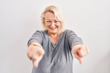 Beyaz arka planda duran orta yaşlı beyaz kadın seni ve kamerayı işaret ediyor, gülümsüyor olumlu ve neşeli. 