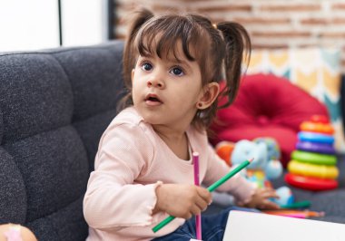 Adorable hispanic girl drawing on notebook sitting on sofa at home