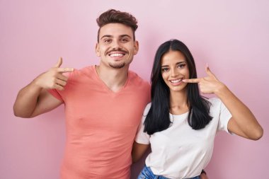 Pembe arka planda duran genç İspanyol çift mutlu bir şekilde gülümseyip parmaklarını ve ağzını işaret ediyor. diş sağlığı kavramı. 