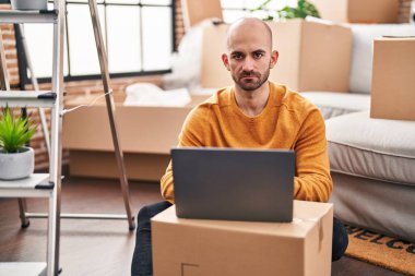 Sakallı genç bir adam dizüstü bilgisayarla yeni bir eve taşınıyor. Düşünme tavrı takınıyor ve kendine güveni tam görünüyor. 