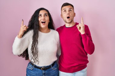 Pembe arka planda duran genç İspanyol çift şaşkınlık ve şaşkınlık içinde başlarını kaldırıp parmaklarını gösterip kollarını kaldırdılar.. 