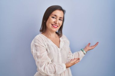 Orta yaşlı Latin bir kadın mavi arka planda durup gülümseyerek doğal bir şekilde içeri girmeye davet ediyor. 