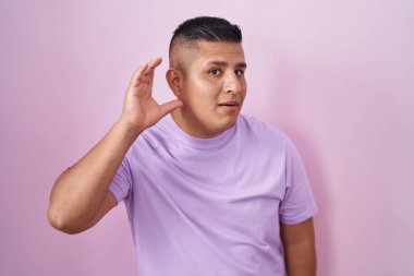 Young hispanic man standing over pink background smiling with hand over ear listening an hearing to rumor or gossip. deafness concept. 