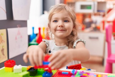 Adorable blonde girl playing with construction blocks sitting on table at kindergarten