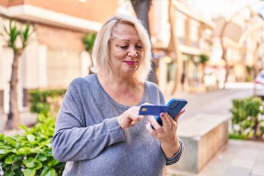 Middle age blonde woman using smartphone and credit card at street