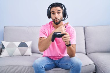 Kanepede oturmuş video oyunu oynayan İspanyol genç adam şaşkın şaşkın parmağını gösterip ağzı açık şaşkın bir ifade sergiliyor.. 