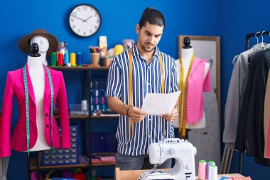 Young hispanic man tailor smiling confident reading document at sewing studio