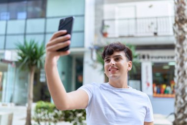 İkili olmayan adam gülümsüyor. Kendine güveni tam. Sokakta akıllı telefonun yanında selfie çekiyor.