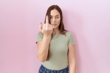 Beautiful brunette woman standing over pink background showing middle finger, impolite and rude fuck off expression 