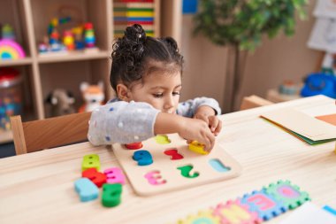 Adorable hispanic girl playing with maths puzzle game sitting on table at kindergarten