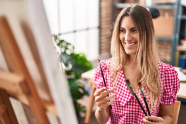 Young woman artist smiling confident holding paintbrushes at art studio