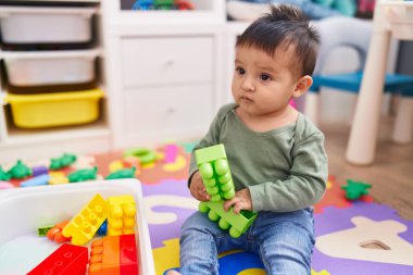 Adorable hispanic boy playing with construction blocks sitting on floor at kindergarten