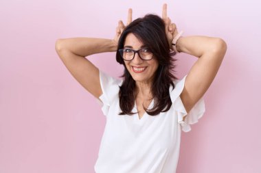 Orta yaşlı İspanyol kadın, günlük beyaz tişört ve gözlük takıyor komik ve deli pozu veriyor parmakları tavşan kulağı gibi, neşeli gülümsüyor. 