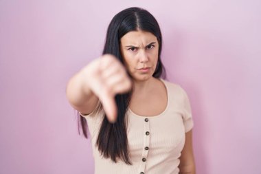 Pembe arka planda duran Latin kökenli genç bir kadın mutsuz ve kızgın görünüyor. Reddedilmiş ve olumsuz bir hareket sergiliyor. kötü ifade. 
