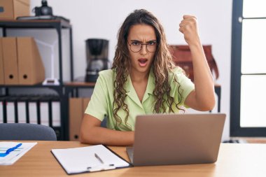 Ofiste çalışan kızgın ve kızgın gözlüklü genç İspanyol bir kadın öfkeyle bağırırken yumruğunu kaldırıyor. öfke ve agresif konsept. 