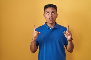 Sarı arka planda dikilen İspanyol genç adam şaşırmış ve şaşırmış bir şekilde yukarı bakıyor ve parmaklarıyla işaret ediyor ve kollarını kaldırıyor.. 