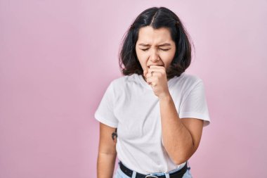 Pembe arka plan üzerine günlük beyaz tişört giyen genç İspanyol kadın kendini iyi hissetmiyor ve soğuk algınlığı veya bronşitin belirtisi olarak öksürüyor. sağlık hizmeti kavramı. 