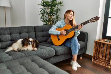 Klasik gitar çalan beyaz bir kadın evdeki kanepede köpeğiyle oturuyor.