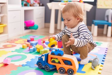 Beyaz tenli sevimli bir çocuk anaokulunda yerde oturmuş oyuncaklarla oynuyor.