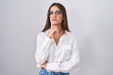 Genç esmer kadın gözlük takıyor şüpheye odaklanmış, parmakları çenesinde ve yukarı bakıyor merak ederek. 