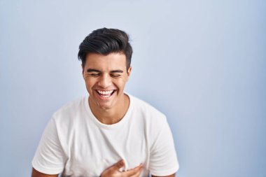 Hispanic man standing over blue background smiling and laughing hard out loud because funny crazy joke with hands on body. 