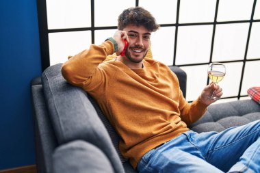 Young hispanic man talking on the smartphone and drinking wine at home