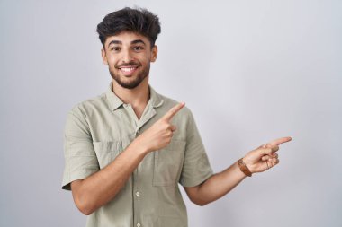 Beyaz arka planda duran sakallı Arap adam gülümsüyor ve kameraya bakıyor iki eliyle ve parmaklarıyla kenarı işaret ediyor.. 