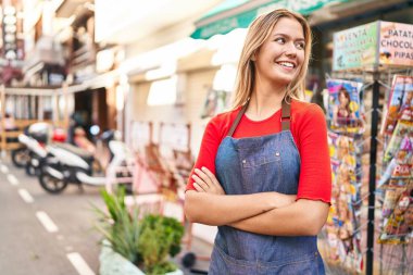 Young hispanic woman shop assistant standing with arms crossed gesture at street