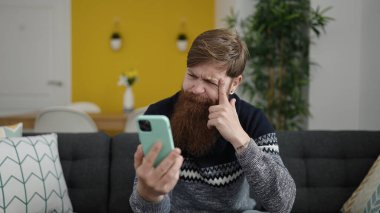 Young redhead man using smartphone sitting on sofa at home