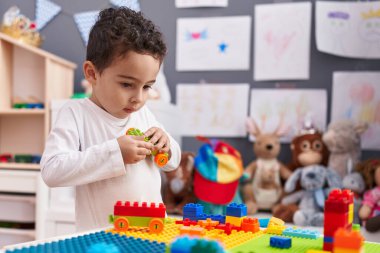 Adorable hispanic boy playing with construction blocks standing at kindergarten