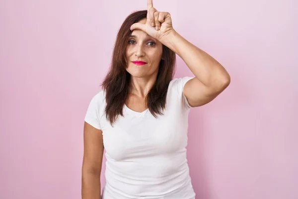 中年黑发女人站在粉红的背景上取笑额头上的人做失败者的手势嘲笑和侮辱 — 图库照片