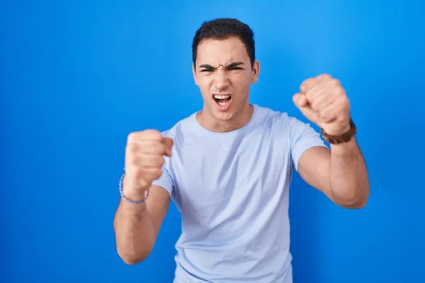 青い背景に立っている若いヒスパニック系の男は怒りで叫んでいる間に怒りと怒りの拳を上げ怒っている 怒りと攻撃的な考え方 — ストック写真