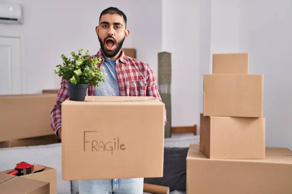 Middle east man with beard moving to a new home holding cardboard box in shock face, looking skeptical and sarcastic, surprised with open mouth