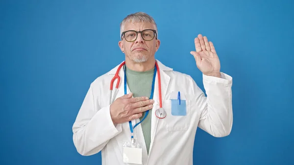 中年白发苍苍的男医生 手托着胸膛在孤零零的蓝色背景下宣誓 — 图库照片
