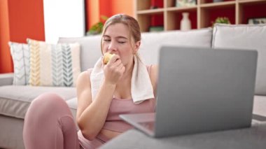 Evde dizüstü bilgisayar kullanarak elma yiyen genç sarışın kadın.