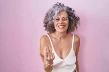 Middle age woman with grey hair standing over pink background beckoning come here gesture with hand inviting welcoming happy and smiling 