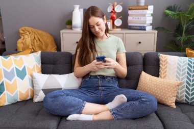 Evdeki kanepede oturan akıllı telefon kullanan genç ve güzel bir kadın.