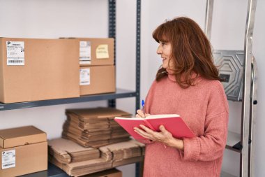 Middle age woman ecommerce business worker writing on book at office