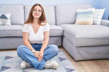 Genç beyaz kadın oturma odasında yerde oturuyor gülümsüyor yan tarafa bakıyor ve düşünüyor.. 