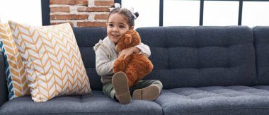 Adorable hispanic girl hugging teddy bear sitting on sofa at home