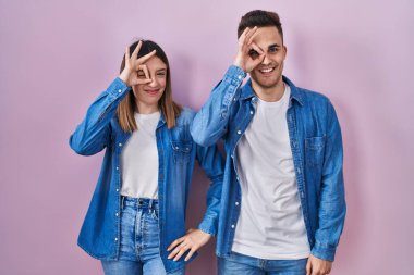 Young hispanic couple standing over pink background doing ok gesture with hand smiling, eye looking through fingers with happy face. 