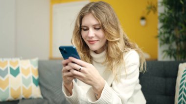 Evdeki kanepede oturan akıllı telefon kullanan genç sarışın kadın.