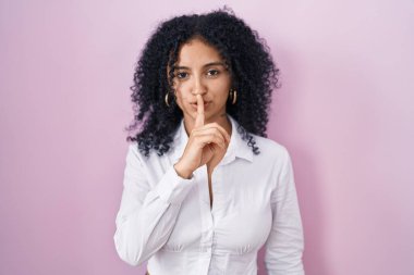 Pembe arka planda duran kıvırcık saçlı İspanyol kadın sessiz olmak istiyor. sessizlik ve gizli konsept. 