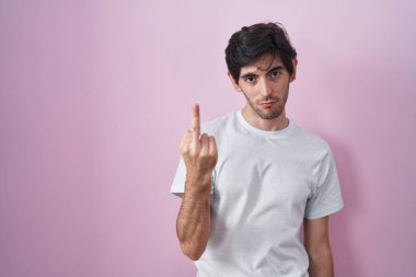 Young hispanic man standing over pink background showing middle finger, impolite and rude fuck off expression 