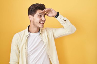 Young hispanic man standing over yellow background very happy and smiling looking far away with hand over head. searching concept. 