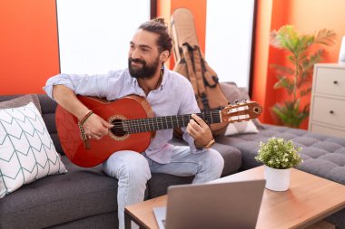 Young hispanic man having online guitar class sitting on sofa at home