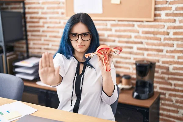 Young girl with blue hair holding model of female genital organ at the office complaining for menstruation pain with open hand doing stop sign with serious and confident expression, defense gesture